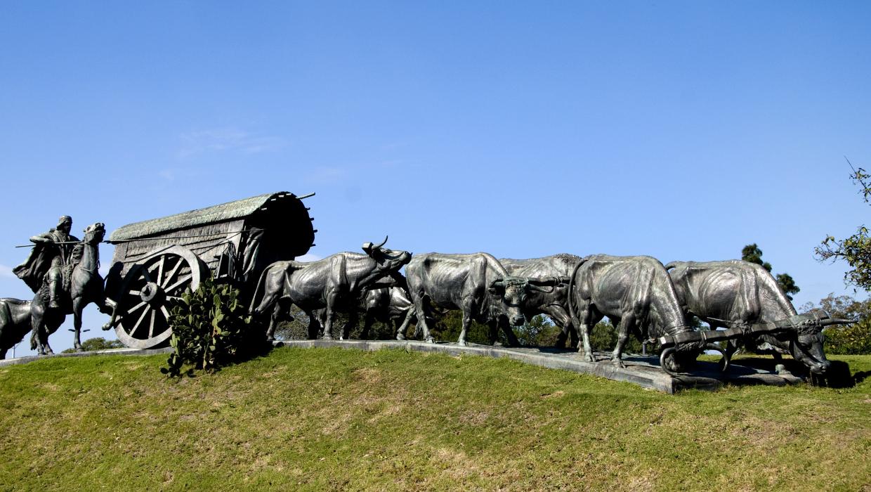 Monumento de una una carreta, con tres yuntas de bueyes tirando de ella y dos bueyes en la parte posterior. Delante de estos últimos, a la derecha del vehículo, un gaucho a caballo empuña la picana. El conjunto, en la parte alta de un terraplén cubierto de césped de tal manera que da la sensación de que avanza por el campo, dominando un estanque que le pone distancia al observador.