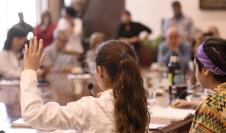 Varios niños con túnica en una mesa redonda con micrófono, levantando la mano y participando. Hay algunos adultos en segundo plano observandolos.