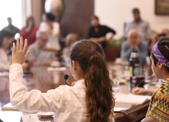 Gabinete recibe al parlamento de niñas, niños y adolescentes Varios niños con túnica en una mesa redonda con micrófono, levantando la mano y participando. Hay algunos adultos en segundo plano observandolos.